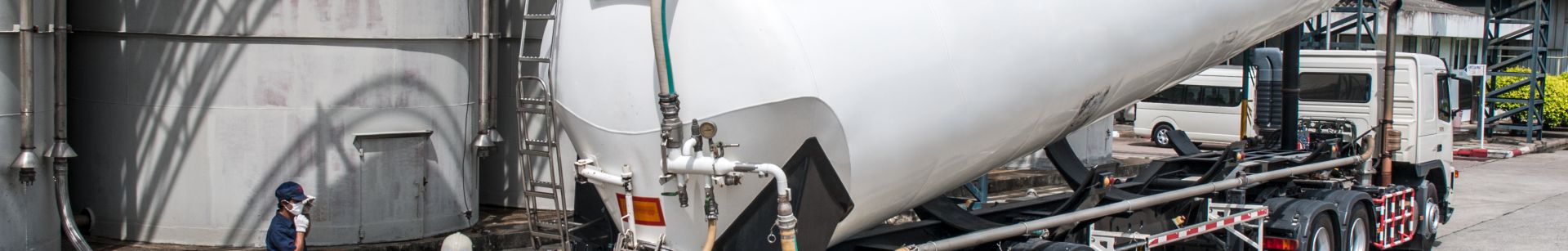 Two workers in safety gear monitor the unloading of a large white tanker truck at an industrial facility