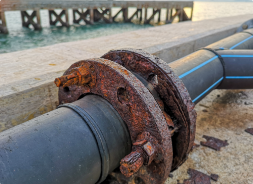 A close-up of a rusted metal flange connecting two black pipes, with a pier and water in the background