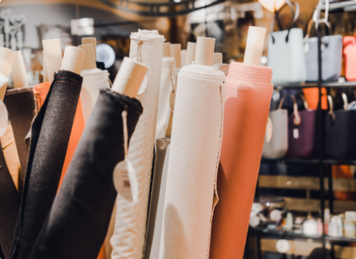 Rolls of fabric in various colors and textures displayed in a store, with shelves of bags and accessories in the background